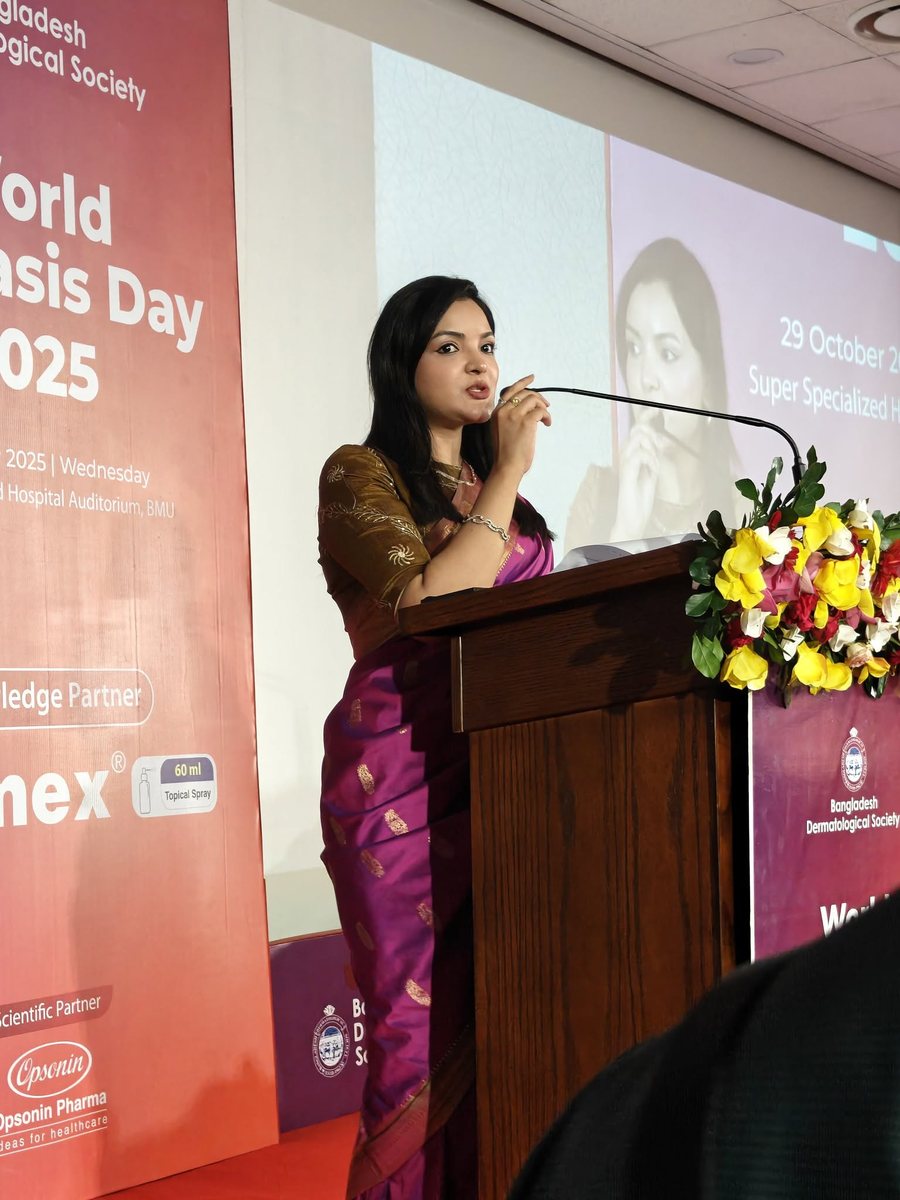 Dr. Tasmin Tahnin Siha speaking at a podium at a World Psoriasis Day event held by the Bangladesh Dermatological Society, with the event banner and an audience screen behind her.